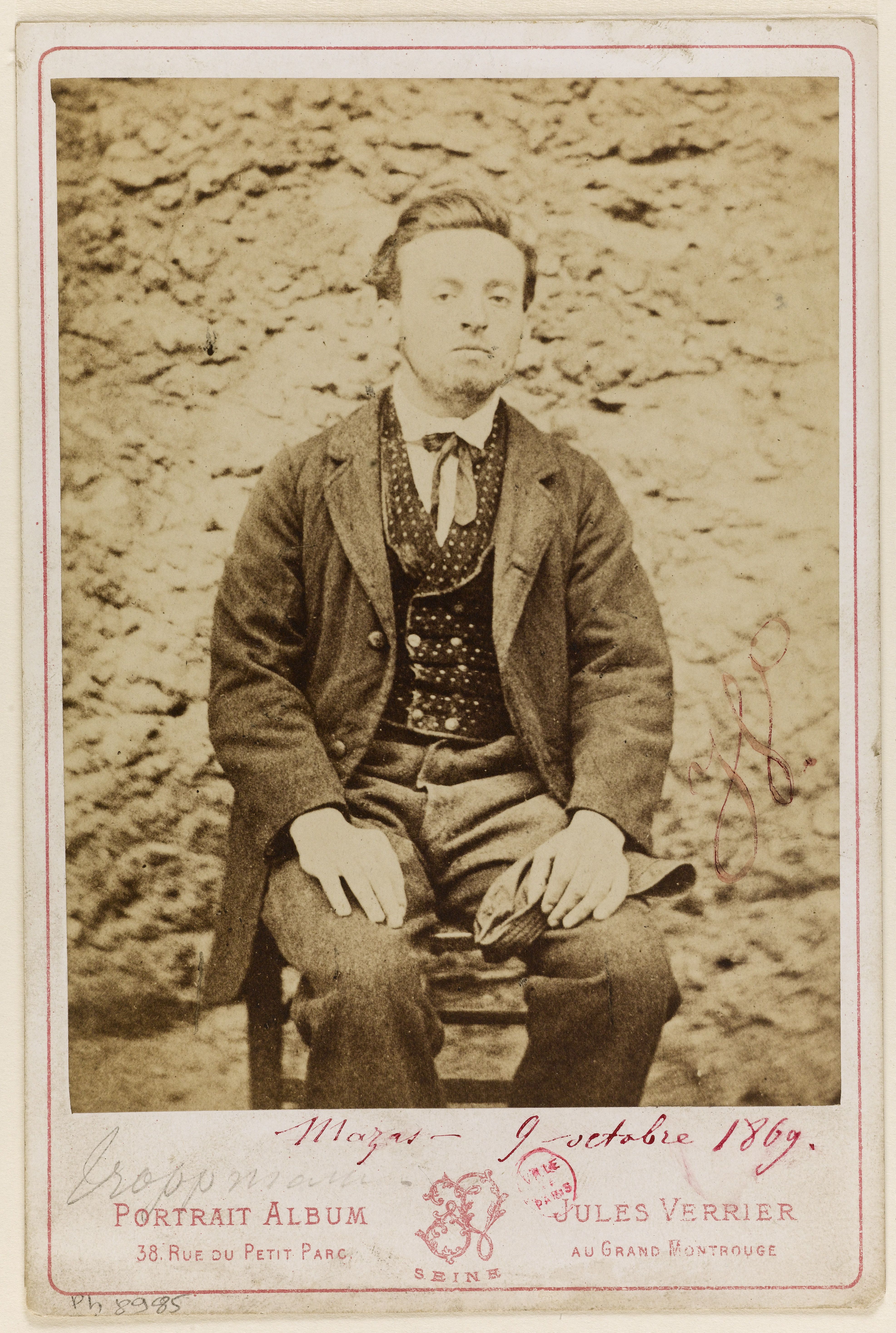 Photographie d'un jeune homme assis devant un mur, en veste et gilet à pois, une casquette à la main ; le montage cartonné porte l'inscription manuscrite « Mazas – 9 octobre 1869 » et la mention imprimée « Portrait Album – Jules Verrier ».