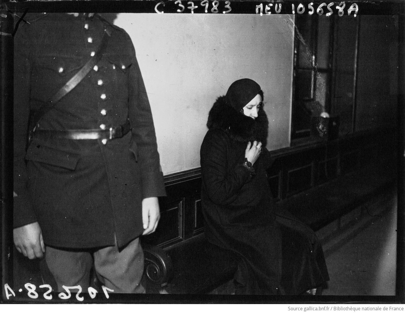 Photographie de presse de 1933 : Violette Nozière, assise sur un banc dans un couloir, le col de son manteau relevé, à côté d'un garde en uniforme.